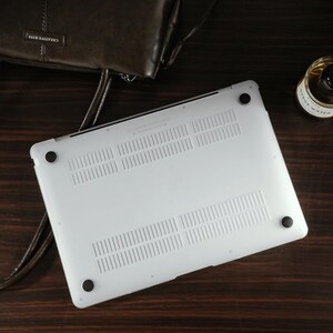 May include: A white laptop protective case featuring a series of vertical ventilation slots and four circular black rubber feet on the underside. The case is positioned on a dark surface next to a brown leather bag and a small glass bottle.