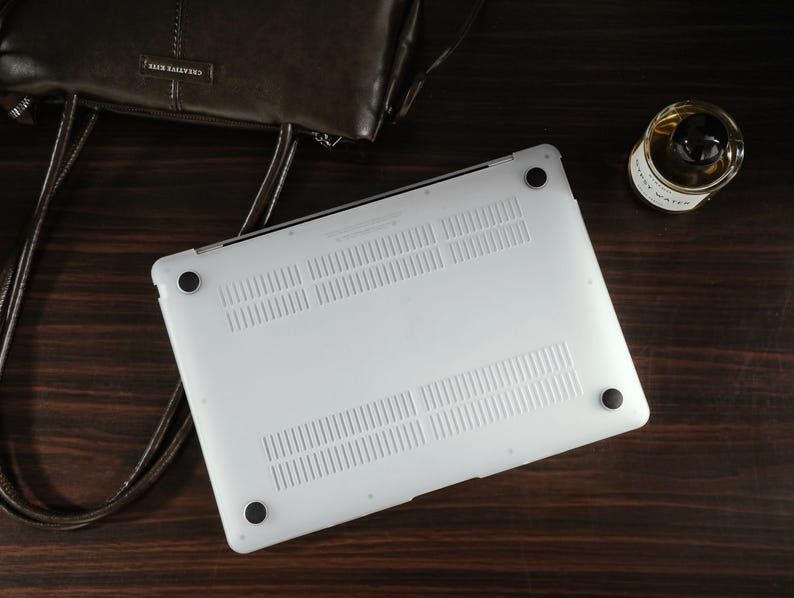 May include: A white laptop protective case featuring a series of vertical ventilation slots and four circular black rubber feet on the underside. The case is positioned on a dark surface next to a brown leather bag and a small glass bottle.