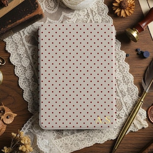 May include: A notebook with a beige cover and a pattern of small red polka dots. The initials "A.S" are in gold on the lower right corner. The notebook is on a wooden surface with a lace doily, a pocket watch, and a quill pen.