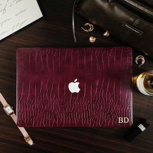 May include: A deep burgundy laptop cover with a textured crocodile skin pattern and a cutout for the Apple logo. The cover features the gold-colored initials BD printed in the bottom right corner, providing a personalized touch to the device.