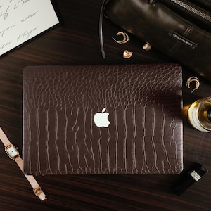 May include: A dark brown laptop cover with a textured crocodile skin pattern featuring a central cutout for the white Apple logo. The rectangular cover is designed to fit over the lid of a laptop, showcasing a deep brown color and embossed finish.