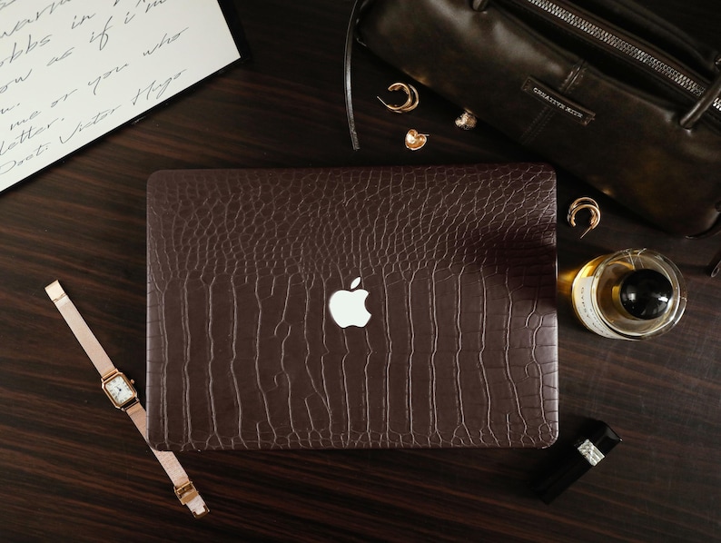 May include: A dark brown laptop cover with a textured crocodile skin pattern featuring a central cutout for the white Apple logo. The rectangular cover is designed to fit over the lid of a laptop, showcasing a deep brown color and embossed finish.