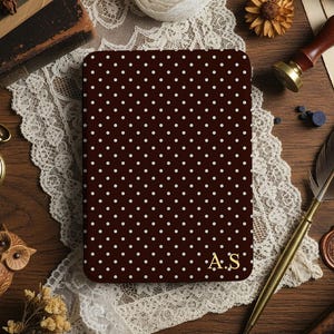 May include: A dark brown notebook with a white polka dot pattern and gold initials "A.S." is displayed on a wooden surface. The notebook is surrounded by vintage items, including a pocket watch, quill, and lace.