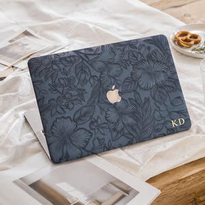 May include: A dark blue laptop cover featuring an embossed floral pattern of hibiscus flowers and leaves. The cover includes a central cutout for the Apple logo and the gold-colored initials KD printed in the bottom right corner of the surface.