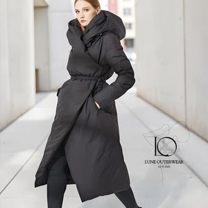 May include: A long, black wrap coat with a large hood and a waist tie. The coat is made of a puffy material and has pockets. The Lune Outerwear logo is visible on the lower right side of the image.