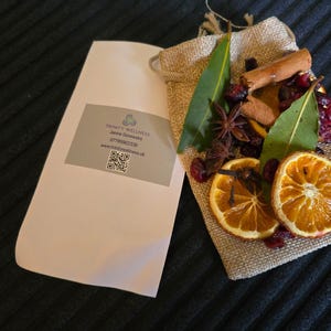 May include: A hessian bag containing dried orange slices, cinnamon sticks, star anise, and bay leaves. A white paper packet with a printed label and QR code is next to the bag. The label reads "Trinity Wellness".
