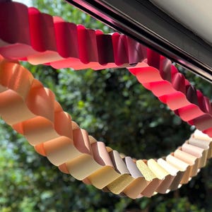 May include: A decorative paper chain garland featuring a gradient of colors ranging from deep burgundy and bright red to soft peach, beige, and cream. The chain is constructed from interlocking paper loops, creating a continuous wavy, scalloped form.