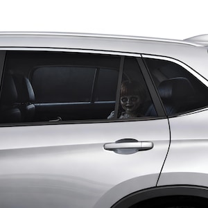 Op de afbeelding: Een zilverkleurige auto met een donker getint achterzijruitje. Een pop met bruin haar en een bleek gezicht is door het raam zichtbaar op de achterbank. De auto heeft een zilveren deurklink en een zwart raamkozijn tegen een witte achtergrond.