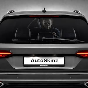 Op de afbeelding: Een grijze stationwagen met een achterruitsticker van een gemaskerd figuur met een mes. Het voertuig heeft rode led-achterlichten, een achterruitenwisser en een kentekenplaathouder met de tekst AutoSkinz in het zwart op wit.