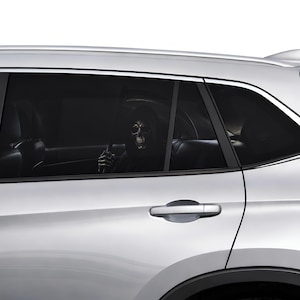 Op de afbeelding: Een zilverkleurige auto met een donker getint achterruitje waarop een afbeelding van een grimmige maaier met een zeis te zien is. De figuur is zichtbaar door het glas, met een skeletachtig gezicht en een donkere kapmantel in de auto.