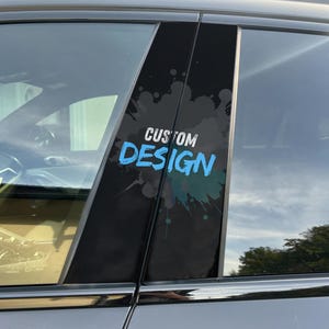 Op de afbeelding: Een zwarte autoraampilaar met de woorden "CUSTOM DESIGN" in wit en blauw, met een teal en grijs spatterontwerp. De autoruit is zichtbaar op de achtergrond.