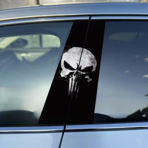 May include: Black car window sticker with a white skull design. The skull has a distressed, textured look with noticeable eye sockets and a vertical line down the middle. The sticker is applied to the car's window frame.