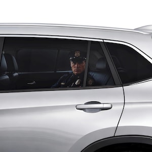 Op de afbeelding: Een zilverkleurige auto met donker getinte achterruiten met een zichtbare afbeelding van een politieagent in uniform met een pet. De sticker is aan de buitenkant van het raam aangebracht, waardoor het lijkt alsof er iemand in de auto zit.