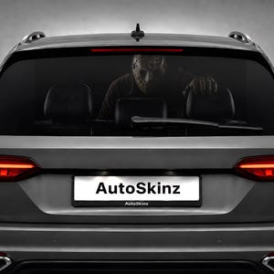 May include: A gray station wagon featuring a rear window decal of a hockey-masked figure peering from inside the vehicle. The car has red LED taillights, a rear wiper, and a license plate frame displaying the text AutoSkinz in black lettering.