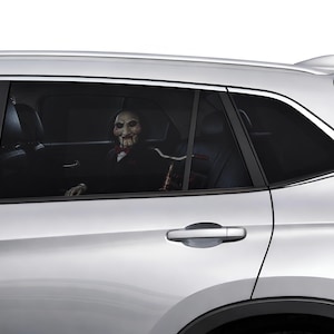 Puede incluir: Un coche plateado con una ventana trasera tintada que muestra una calcomanía de un muñeco ventrílocuo con cara blanca, pelo negro, mejillas en espiral rojas y pajarita roja, posicionado como si estuviera sentado en el asiento trasero.