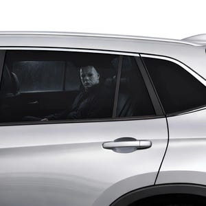 May include: A silver SUV with tinted windows. A person wearing a white mask and black clothing is seated in the back seat. The background shows a rainy, overcast day.