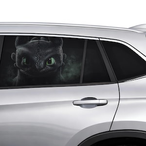 Può includere: Un adesivo per finestrino auto con un primo piano di un drago nero con occhi verde brillante. Il drago è ambientato su uno sfondo scuro e fumoso. L'adesivo è applicato sul lunotto posteriore di un'auto argentata.