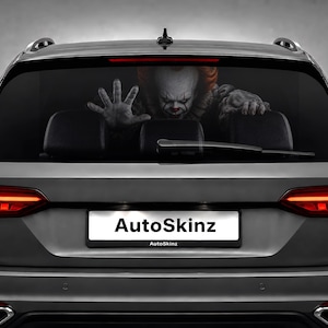 Könnte beinhalten: Ein Auto-Heckscheibenaufkleber mit der Grafik eines Clowns mit roten Haaren und weiß geschminktem Gesicht, der seine Hände gegen die Scheibe drückt. Der Kennzeichenhalter zeigt den Text AutoSkinz in schwarzer Schrift auf weißem Grund.