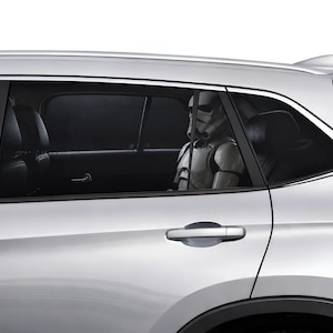 Pode incluir: Um carro prateado com vidros traseiros fumados, onde um capacete de Stormtrooper branco e preto é visível no banco de trás. O veículo tem um puxador da porta na cor da carroçaria, molduras pretas nas janelas e uma antena de barbatana.