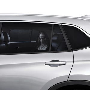 Op de afbeelding: Een zilverkleurige auto met getinte achterruiten waarin een figuur in een traditioneel zwart-wit nonnenhabijt met een bleek gezicht en donkere make-up op de achterbank zit. Het voertuig heeft een zilveren deurklink en een haaienvinantenne.