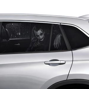 Pode incluir: Um carro prateado com um adesivo na janela apresentando um gráfico em preto e branco de um personagem com pintura facial branca, maquiagem escura nos olhos e jaqueta escura, sentado no banco de trás com a mão no caixilho da janela.