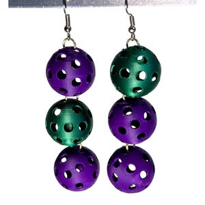May include: A pair of dangle earrings featuring three perforated spheres in alternating purple and green hues. The spheres are connected by silver-colored hooks and wires. The earrings are displayed against a white background.