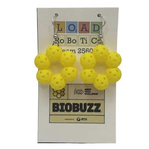 May include: A pair of dangle earrings featuring bright yellow, spherical, perforated pickleball-style beads arranged in a flower shape. The earrings are attached to a white display card printed with the text LOAD ROBOTICS Team 2560, FIRST TECH CHALLENGE, and BIOBUZZ.