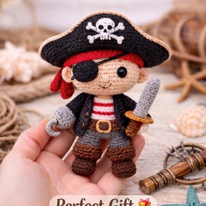 Op de afbeelding: Gehaakte piratenpop met zwarte hoed, rode bandana en ooglap. De pop heeft een haak en een klein zwaard. De tekst "Perfect Gift" is zichtbaar onderaan de afbeelding.