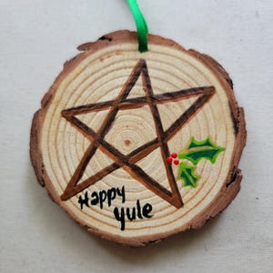 May include: A wooden round ornament with a brown pentagram painted on it. The ornament is decorated with green holly leaves and red berries. The words "Happy Yule" are written in black paint on the wood.