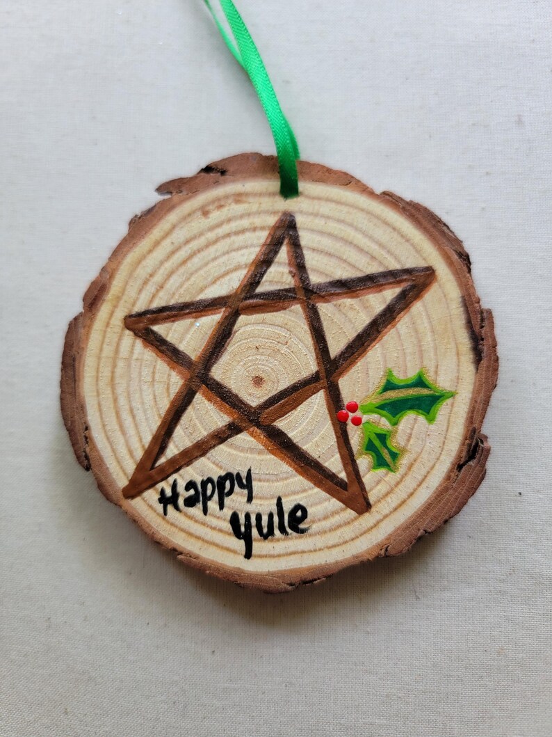 May include: A wooden round ornament with a brown pentagram painted on it. The ornament is decorated with green holly leaves and red berries. The words "Happy Yule" are written in black paint on the wood.