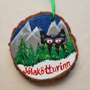 May include: A round wooden ornament with a painted scene of a black cat with red eyes peeking out from behind a snowy mountain range with green pine trees. The text "J&ouml;lak&ouml;tturinn" is written in red on the bottom of the ornament.