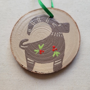 May include: A round wooden ornament with a painted brown goat with green leaves and red berries. The ornament has a green ribbon attached for hanging.