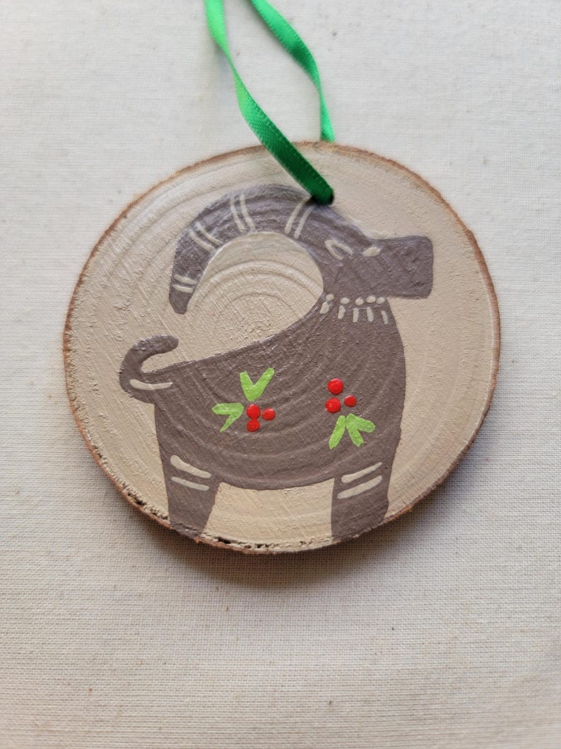 May include: A round wooden ornament with a painted brown goat with green leaves and red berries. The ornament has a green ribbon attached for hanging.