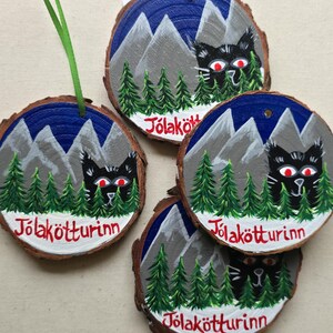 May include: Four round wooden ornaments with a black cat peeking out from behind a forest scene. The ornaments are painted with a blue night sky, gray mountains, green trees, and white snow. Each ornament has the text "J&ouml;lak&ouml;tturinn" written in red on the bottom.