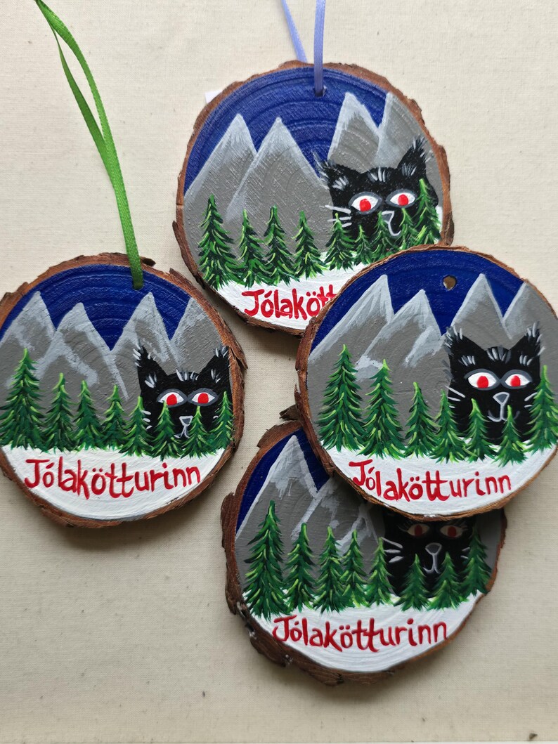 May include: Four round wooden ornaments with a black cat peeking out from behind a forest scene. The ornaments are painted with a blue night sky, gray mountains, green trees, and white snow. Each ornament has the text "J&ouml;lak&ouml;tturinn" written in red on the bottom.