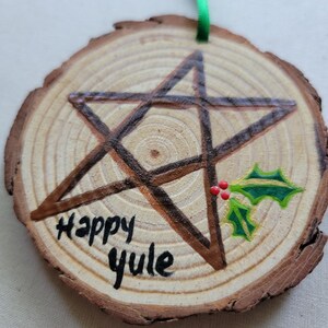 May include: A wooden round ornament with a brown pentagram painted on it. The ornament has the words "Happy Yule" written on it and a green holly sprig with red berries painted on the right side.