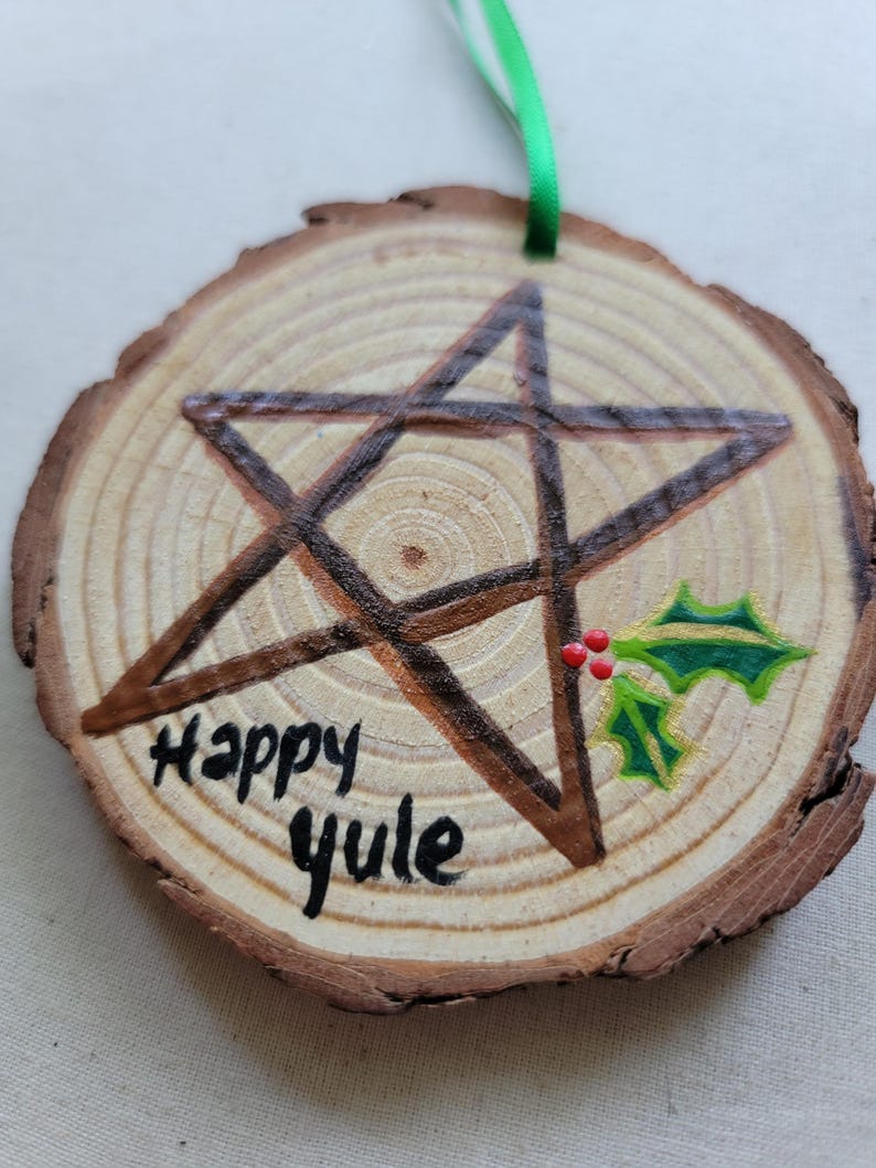 May include: A wooden round ornament with a brown pentagram painted on it. The ornament has the words "Happy Yule" written on it and a green holly sprig with red berries painted on the right side.