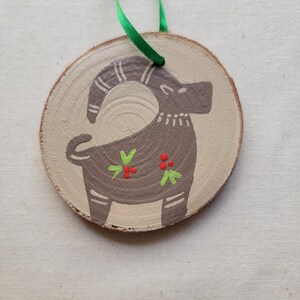 May include: A wooden ornament with a painted brown goat with green and red accents. The ornament is attached to a green ribbon.