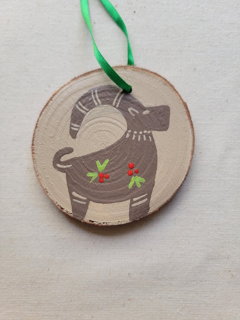 May include: A wooden ornament with a painted brown goat with green and red accents. The ornament is attached to a green ribbon.