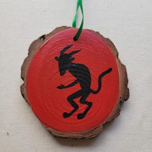 May include: A red wooden ornament with a black silhouette of a goat painted on it. The ornament has a green ribbon attached to it.