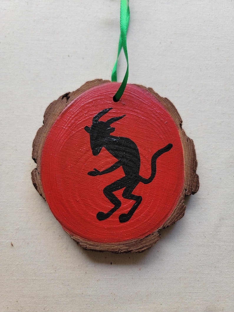 May include: A red wooden ornament with a black silhouette of a goat painted on it. The ornament has a green ribbon attached to it.