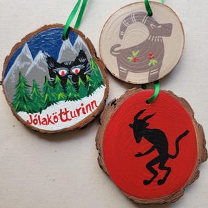 May include: Three round wooden ornaments with painted designs. The first ornament is painted with a black cat in front of a mountain range with a blue sky and the text "J&ouml;lak&ouml;tturinn". The second ornament is painted with a brown goat with red berries. The third ornament is painted with a red background and a black silhouette of a goat.