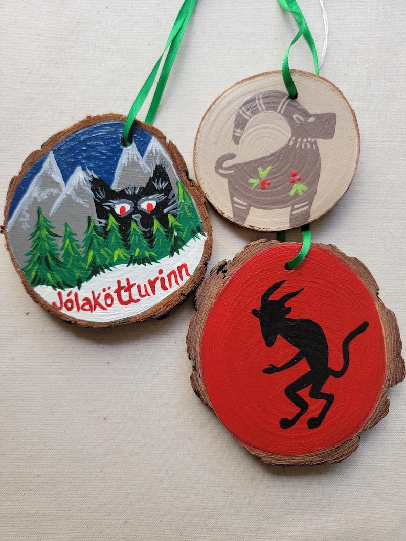 May include: Three round wooden ornaments with painted designs. The first ornament is painted with a black cat in front of a mountain range with a blue sky and the text "J&ouml;lak&ouml;tturinn". The second ornament is painted with a brown goat with red berries. The third ornament is painted with a red background and a black silhouette of a goat.