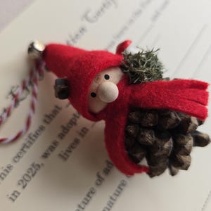 May include: A handmade gnome ornament with a red felt hood and a pinecone body. The gnome has a wooden nose and a small bell attached to a red and white string. The ornament rests on a document with text.