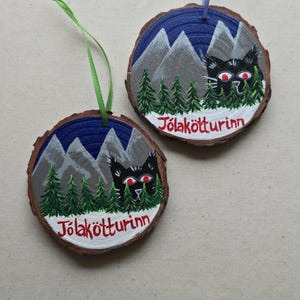 May include: Two round wooden ornaments with a blue and white mountain scene, green trees, and a black cat with red eyes. The text "J&ouml;lak&ouml;tturinn" is written in red on each ornament.