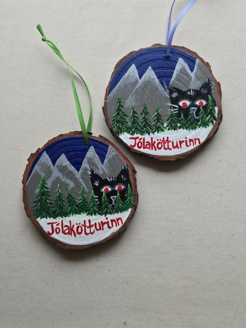 May include: Two round wooden ornaments with a blue and white mountain scene, green trees, and a black cat with red eyes. The text "J&ouml;lak&ouml;tturinn" is written in red on each ornament.