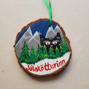 May include: A round wooden ornament with a painted scene of a black cat with red eyes peeking out from behind a snowy forest with mountains in the background. The text "J&ouml;lak&ouml;tturinn" is written in red on the bottom of the ornament.