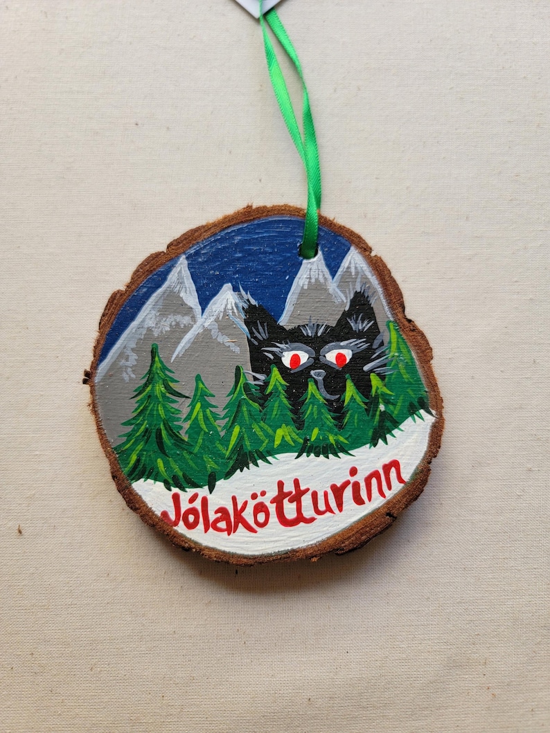 May include: A round wooden ornament with a painted scene of a black cat with red eyes peeking out from behind a snowy forest with mountains in the background. The text "J&ouml;lak&ouml;tturinn" is written in red on the bottom of the ornament.