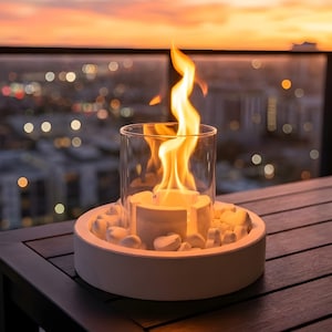 May include: A tabletop fire pit with a bright orange flame inside a clear glass cylinder. The fire pit is white and surrounded by white stones. The background shows a city skyline at sunset.