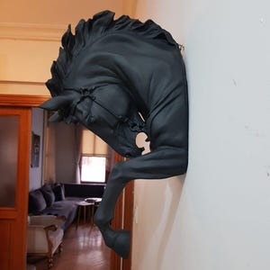 Puede incluir: Escultura de cabeza de caballo negro montada en una pared blanca. La escultura presenta una melena y brida detalladas, con la cabeza y las patas delanteras del caballo que se extienden desde la pared. Una obra de arte que añade elegancia ecuestre.